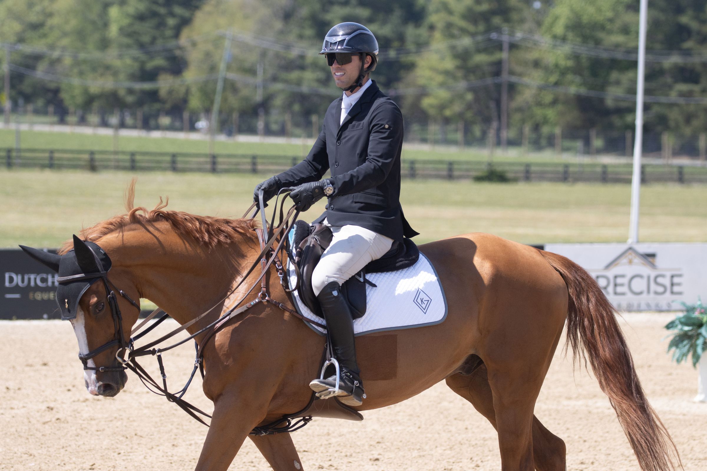 Kyle Gambino riding a chestnut horse in competition attire at a show ring, with the KG diamond logo visible on the saddle pad