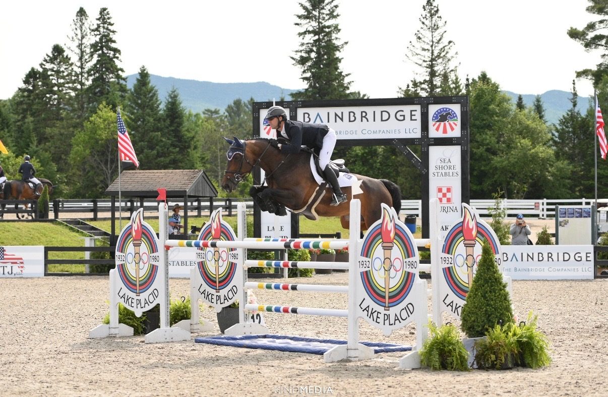 Kyle Gambino on a bay horse mid-jump over an Olympic-themed oxer at Lake Placid, mountains and pine trees in background