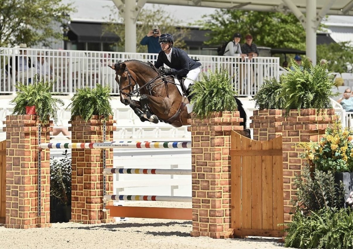 Kyle Gambino mid-jump over a substantial brick-and-wood gate fence at a high-level competition venue, photographers and spectators visible in the background