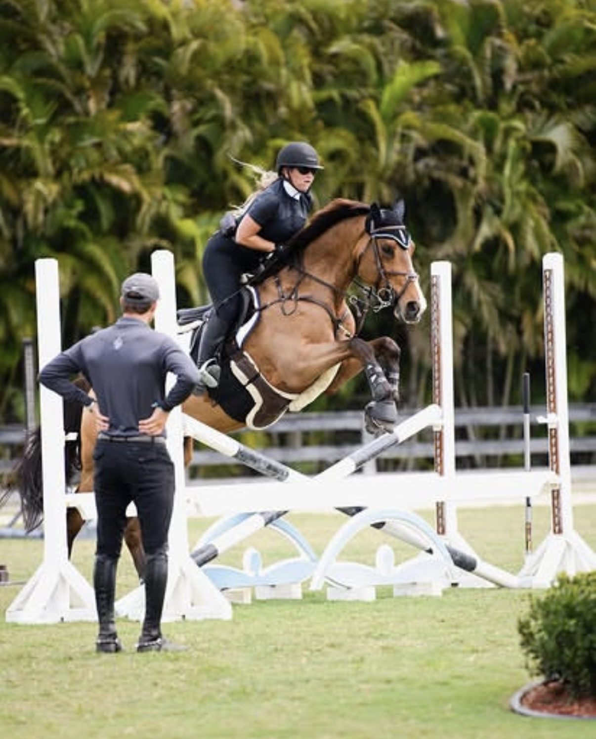 Kyle Gambino in the foreground watching a rider clear a vertical jump in a Florida setting, embodying the coaching relationship between trainer and athlete