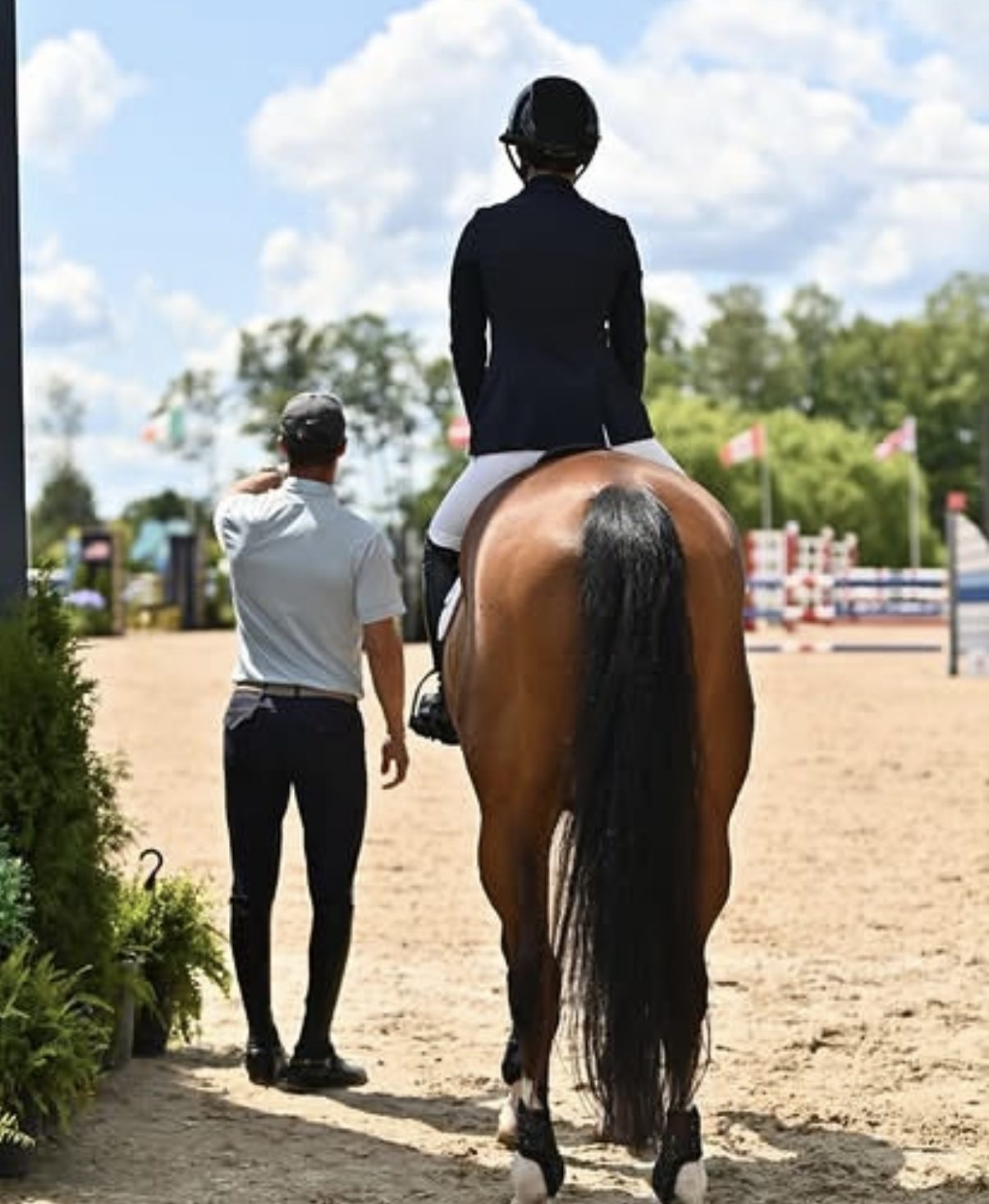 Kyle Gambino and a rider photographed from behind at the in-gate of an international competition, Kyle's hand resting on the rider's back, both looking ahead into the arena