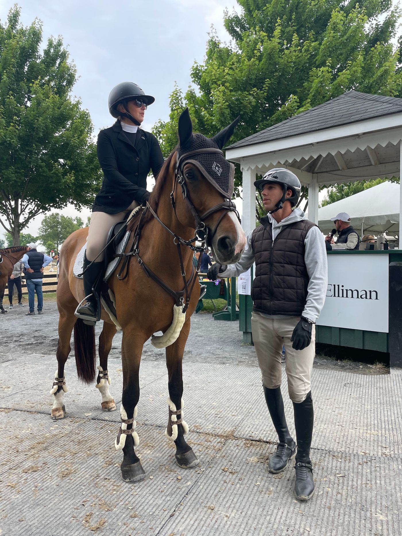 Kyle Gambino standing beside an adult amateur rider mounted on a bay horse, his hand on the horse's neck, both smiling after a good round in golden-hour light