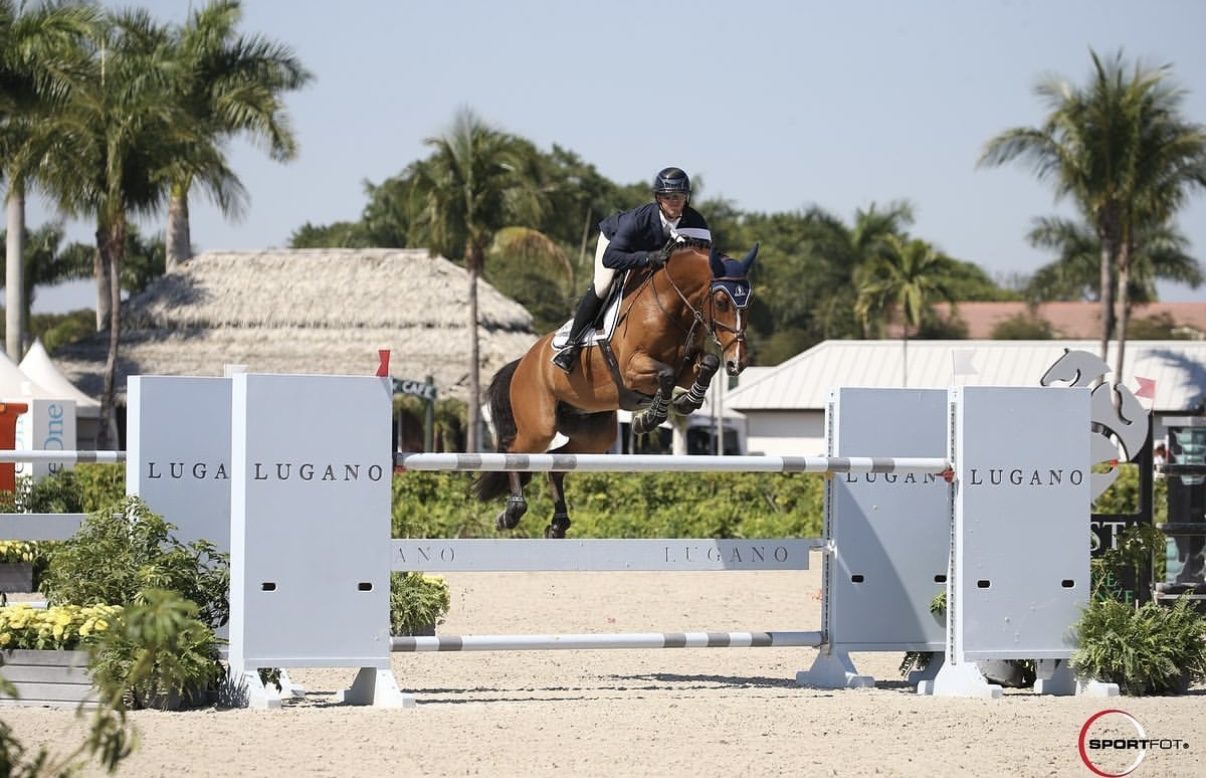 An adult amateur rider on a chestnut horse with Kyle Gambino in coaching position at the in-gate, competition tent visible in the background