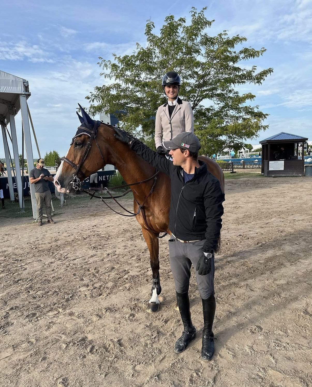 A jumper rider clearing a substantial oxer with Lugano-branded standards at a top-tier Wellington venue, palm trees and competition infrastructure in the background